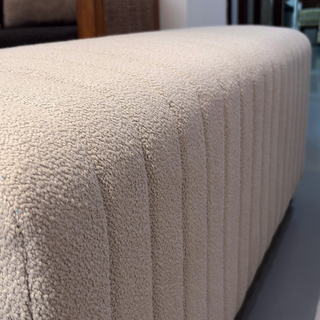 A close-up of a cream-colored bouclé upholstered ottoman or bench with vertical channel tufting, shown in a modern indoor setting.